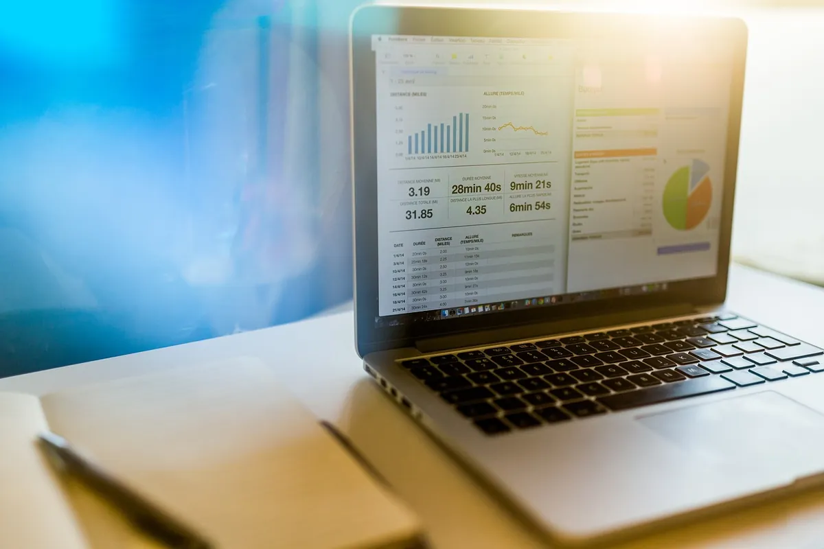 Laptop on a bright desk showing a spreadsheet with a data table and bar and pie charts, next to a notebook and pen