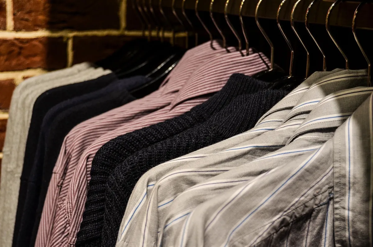 Row of striped shirts and dark sweaters on metal hangers against a brick wall