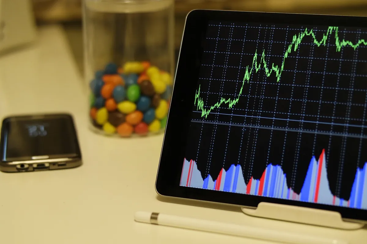 Tablet on a stand showing a rising market chart, next to a phone and a stylus on a desk