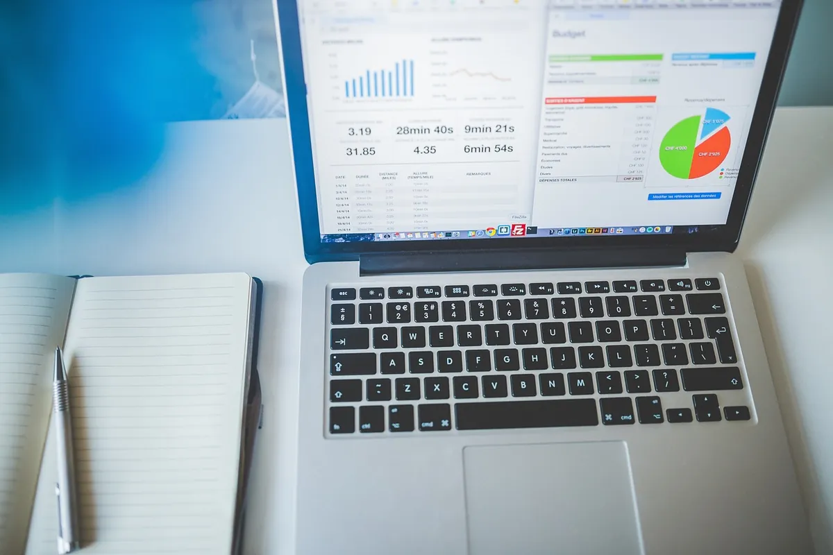 Laptop on a white desk showing a spreadsheet with a data table and charts, next to an open notebook